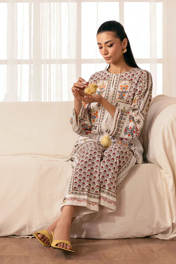 Seated in a sunlit room with soft curtains, printed cambric set in warm tones styled with yellow sandals and two baby chicks, Pomegranate Blossom, Nainpreet.
