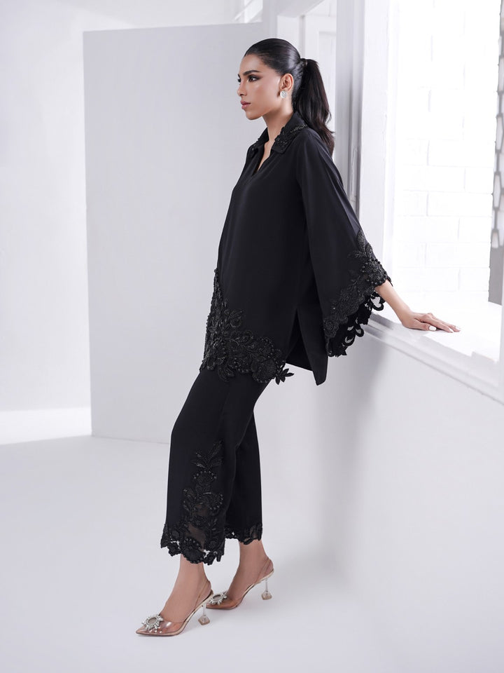 Leaning by the window, the model wears Alvena: a black two-piece with hand-beaded cutwork on the shirt and culottes, styled with heels.