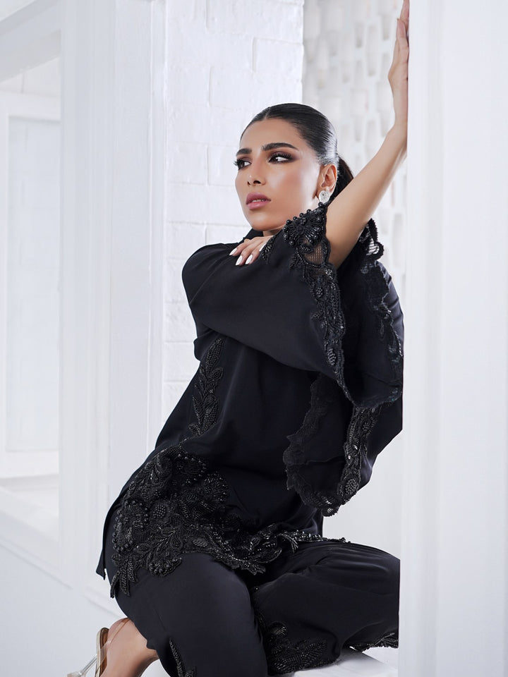 Seated in a modern white space, the model wears Alvena: a black two-piece with hand-beaded appliqué and cutwork on shirt and culottes.