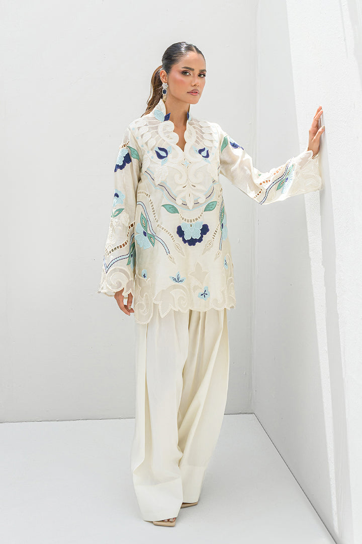 Model showcasing Nadia Farooqui Azure off-white short top with blue floral embroidery and white pleated trousers from Deena Eid Edit '25 collection