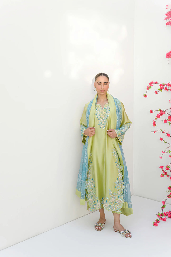 The model stands against a white wall beside a vibrant pink flowering plant, wearing Mint Margarita: a lime-kiwi Sheesha silk tunic with intricate embroidery.