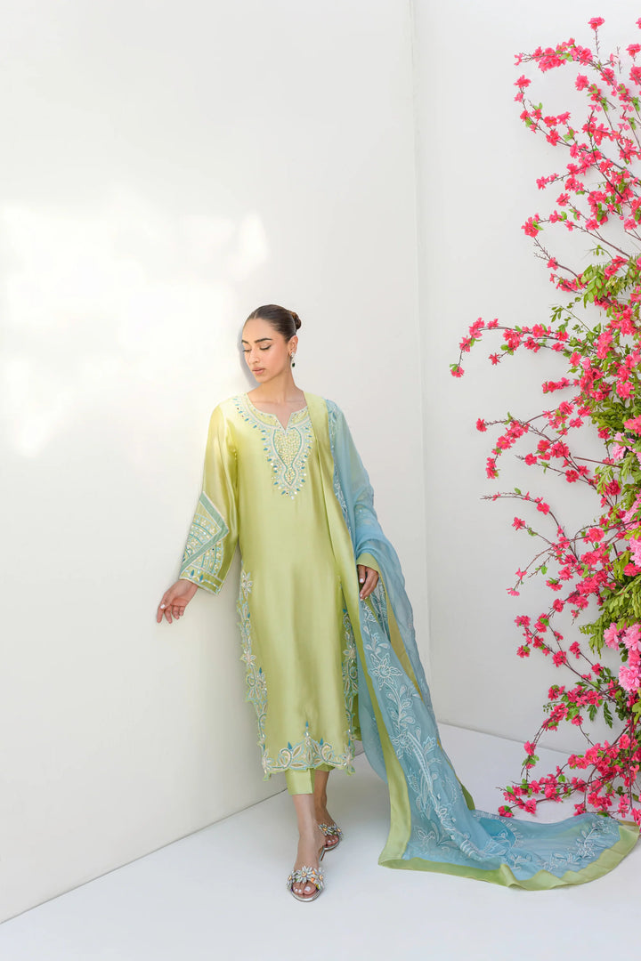 The model stands against a white wall beside a vibrant pink flowering plant, wearing Mint Margarita: a lime-kiwi Sheesha silk tunic with intricate embroidery along the neckline, sleeves, and hem.