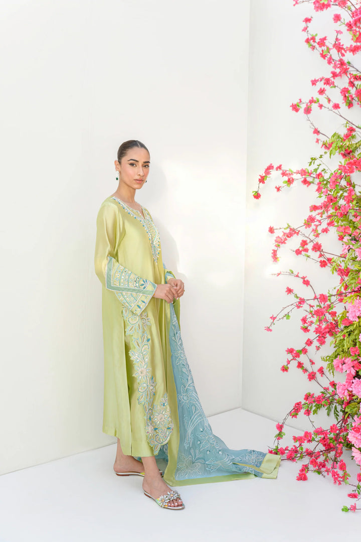 Wearing Mint Margarita from Estelle Eid Edit II’25, the model stands in a minimalist space dressed in a lime-kiwi Sheesha silk shirt with intricate embroidery, sequins, and mirror accents.