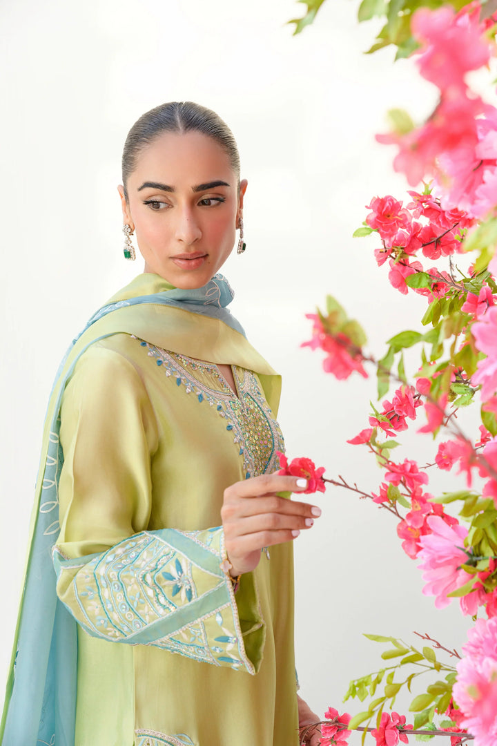 Touching a branch of vibrant pink blossoms, the model wears Mint Margarita: a lime-kiwi Sheesha silk shirt with delicate embroidery, paired with a matching blue and green organza dupatta.