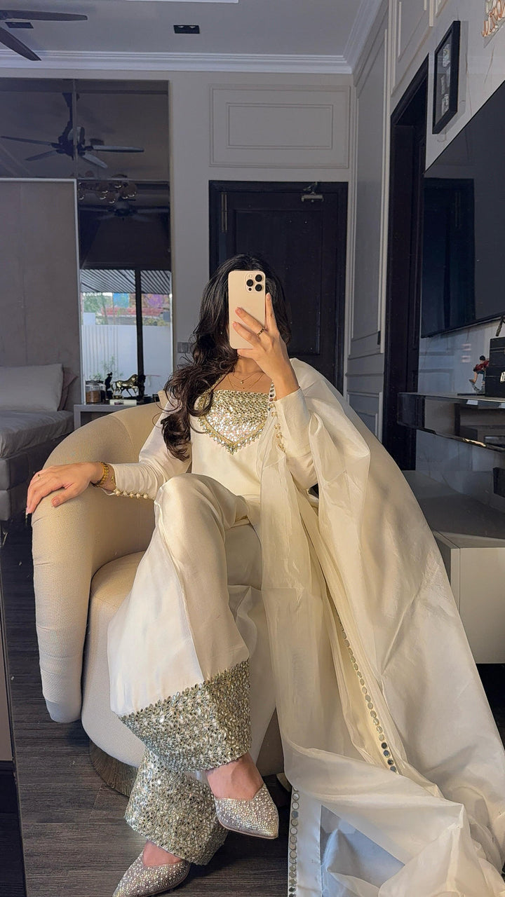 A seated portrait in The Amal Set—ivory silk kurta with mirror-work neckline, farshi shalwar, and shimmer-trimmed dupatta styled casually in a modern room.