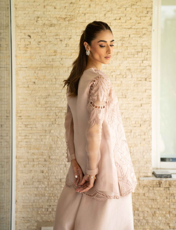 Model stands in Petal Smoke—tea-pink organza jacket with floral cutwork and sheer sleeves, paired with raw silk trousers and soft composure.