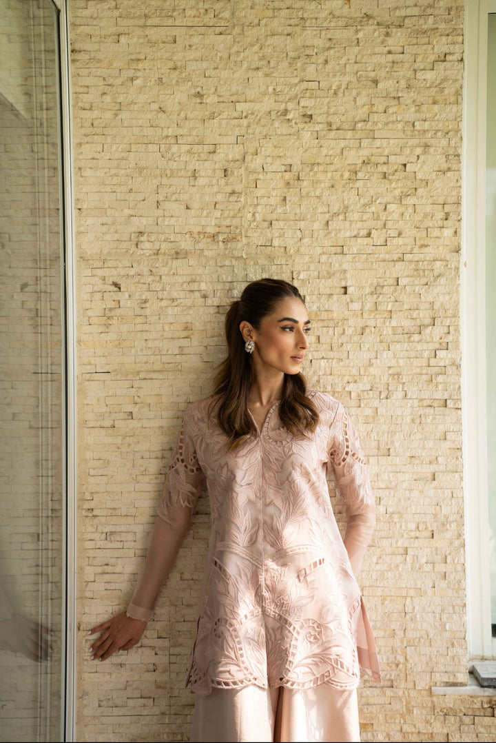 Petal Smoke, worn by the model against stone textures—tea-pink organza jacket with sheer sleeves and cutwork embroidery, paired with raw silk trousers and confidence