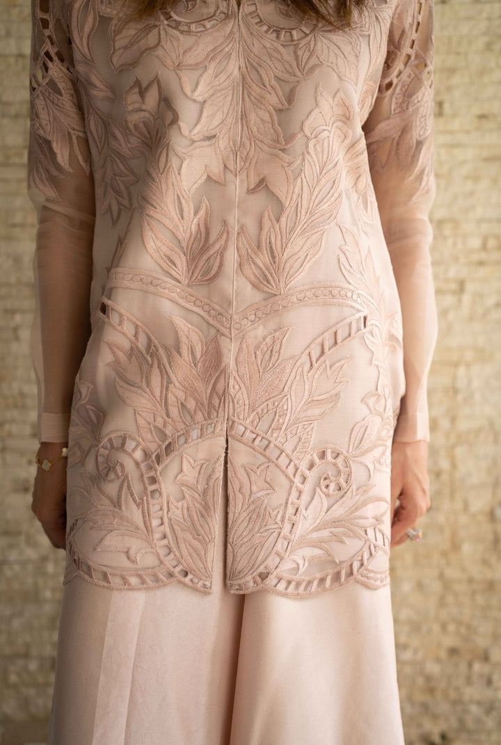 Close-up of Petal Smoke—tea-pink organza jacket with symmetrical floral cutwork, sheer sleeves, and raw silk trousers, captured against stone textures.